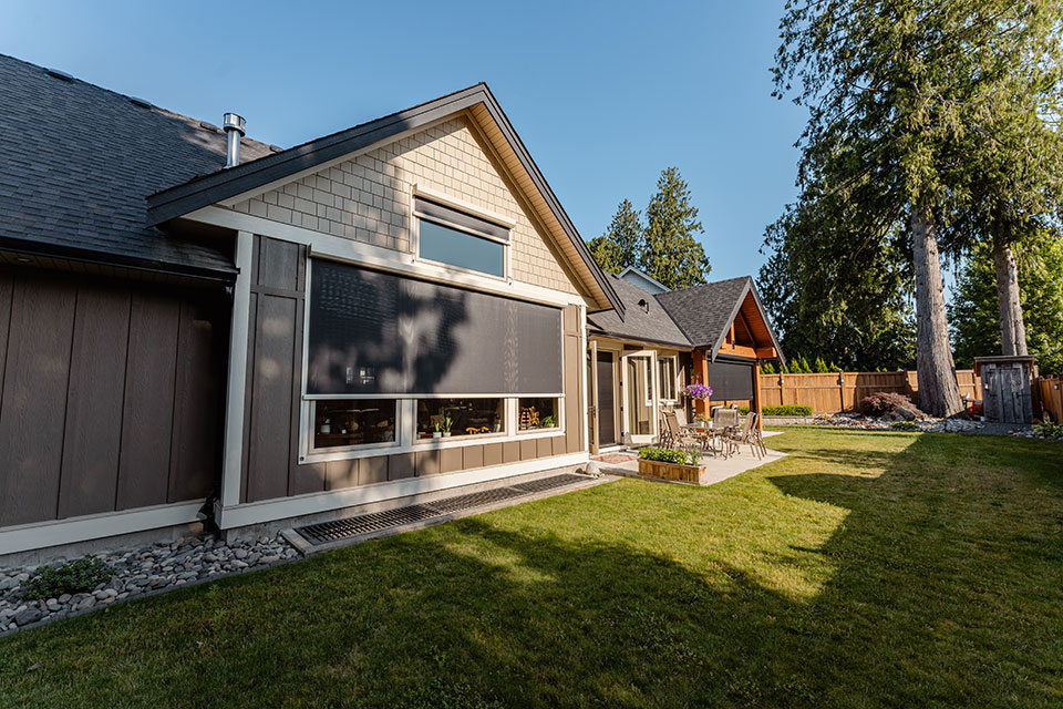 Modern home with exterior retractable screens installed on windows, featuring a landscaped backyard with patio seating and tall trees.