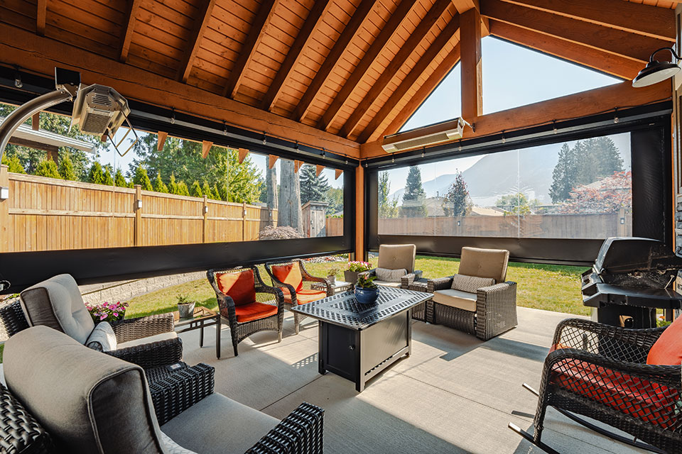 Covered patio with wooden beam ceiling and motorized retractable screens, featuring comfortable outdoor furniture, a fire table, and mountain views in the background.