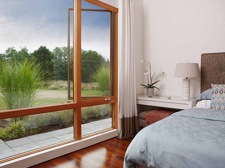 Modern bedroom with large wood-framed window overlooking natural grassland, featuring hardwood floors and elegant bedding